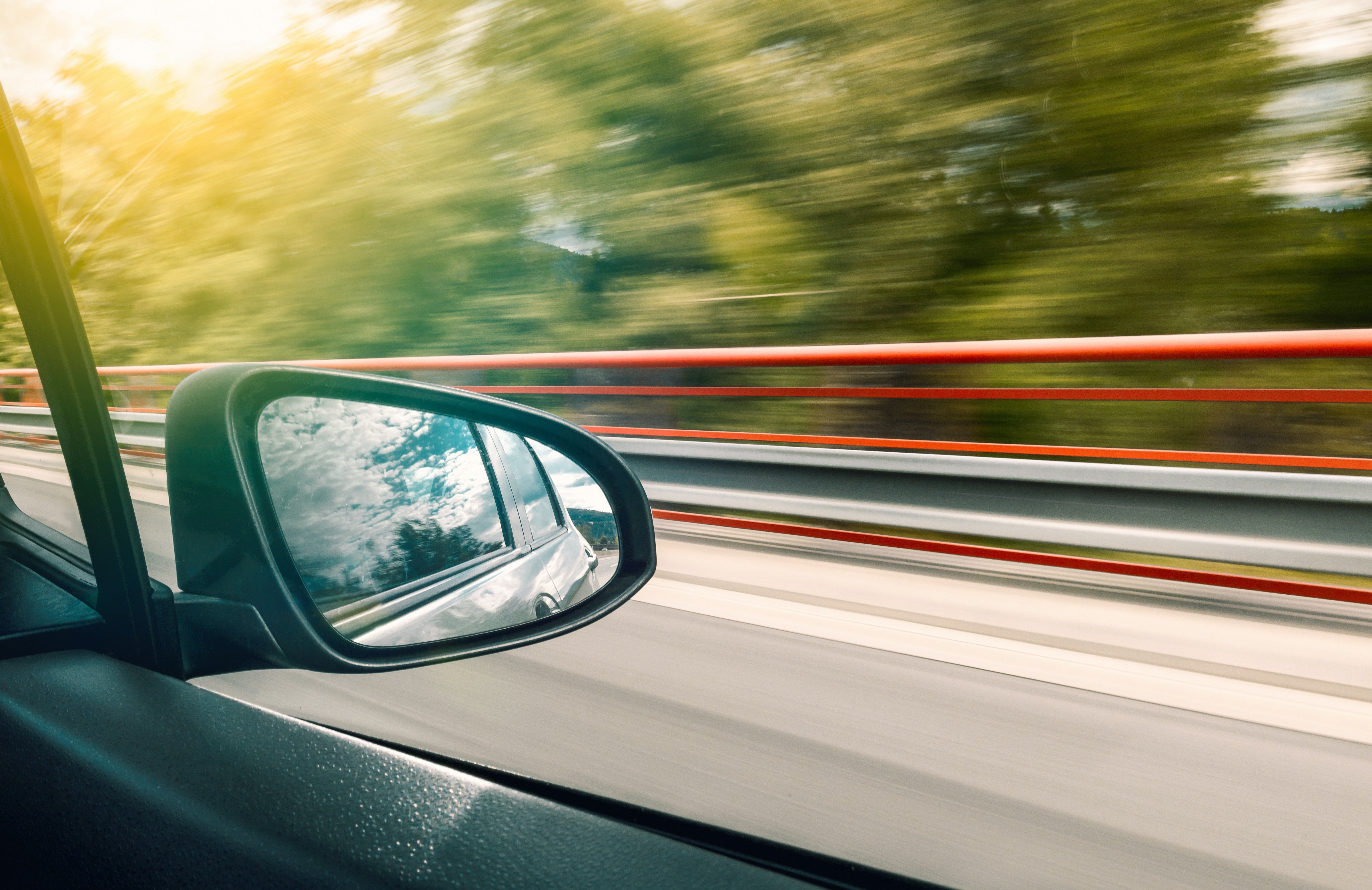 A car's side mirror is shown as the car drives down a forested highway