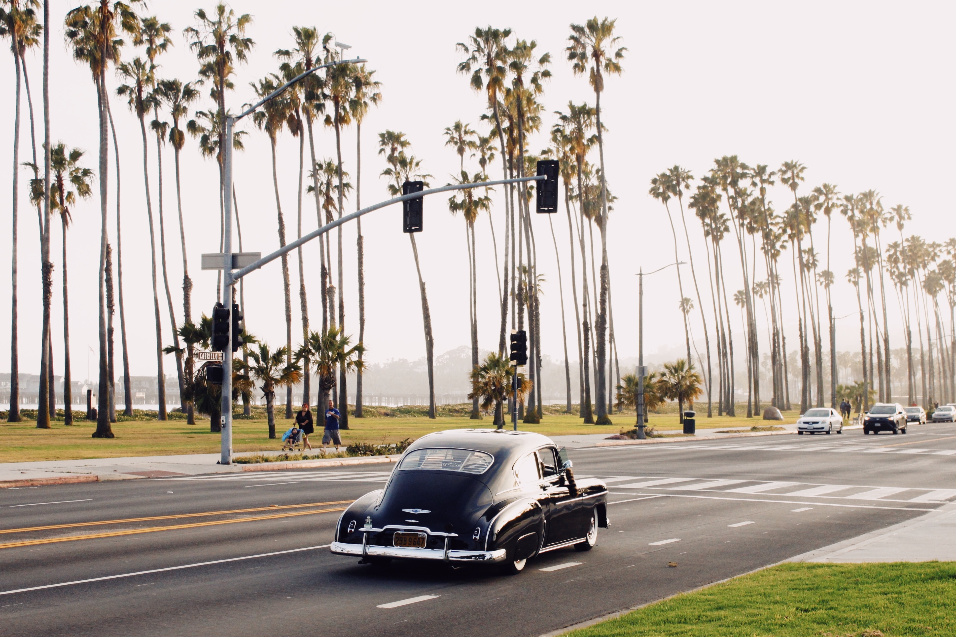 An old-fashioned car drives down a street alongside palm trees and a beach.