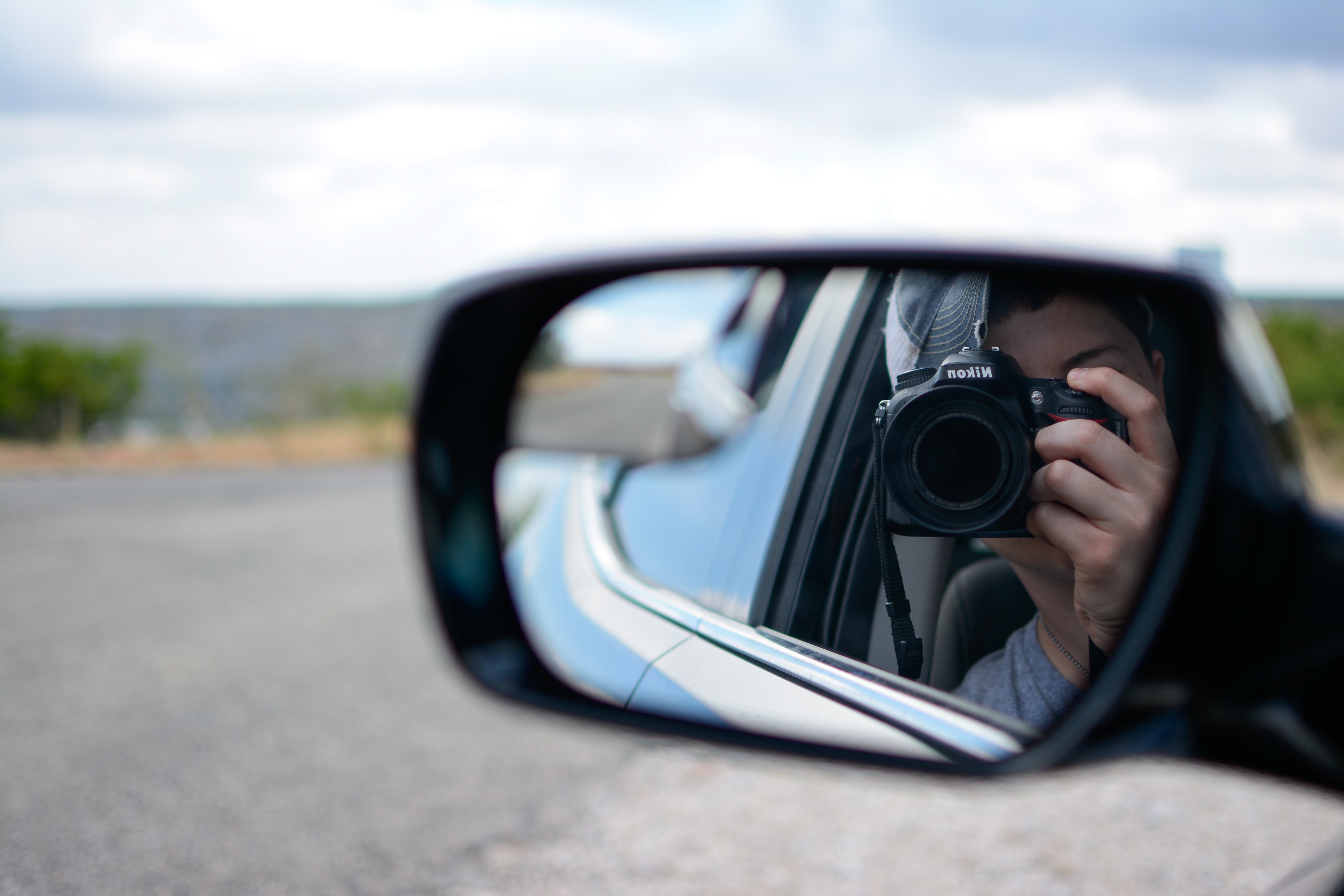 A person seated in a car is reflected in the side mirror of a car, taking a photo of themselves with a Nikon camera.